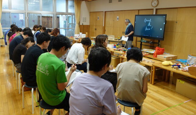 研修会場で講義をする講師と、木製の積木・玩具を前に着席する保育士たち