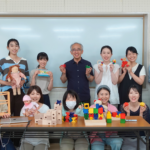 講師を囲み、手作りの積木作品を手に持って笑顔で並ぶ研修参加者の集合写真