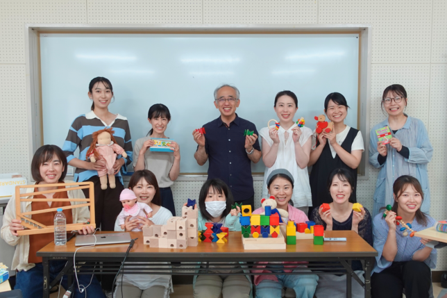 講師を囲み、手作りの積木作品を手に持って笑顔で並ぶ研修参加者の集合写真
