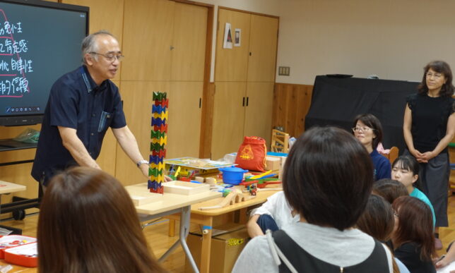 講師が積木を高く積み上げて実演し、参加者が真剣な表情で見入る研修風景