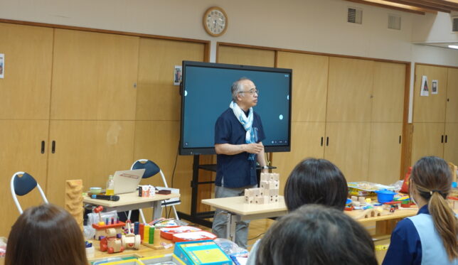研修会場で講義をする講師と、木製の積木・玩具を前に着席する保育士たち
