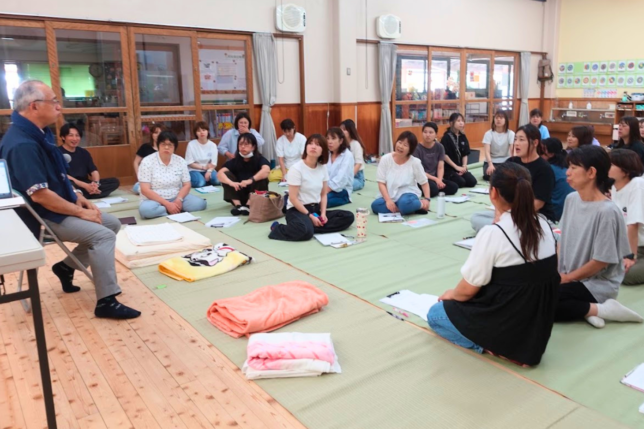 日本知育玩具協会の保育士向け研修風景。講師が椅子に座って講義し、参加者の保育士たちが体育館の床に座って真剣に耳を傾けている様子