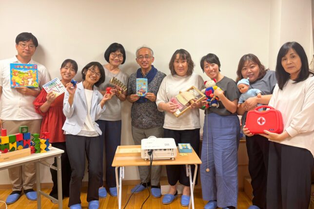保育園の園長先生方を対象とした「おもちゃと子どもの発達」研修の集合写真。木のおもちゃを手にした参加者と講師陣が笑顔で並んでいる