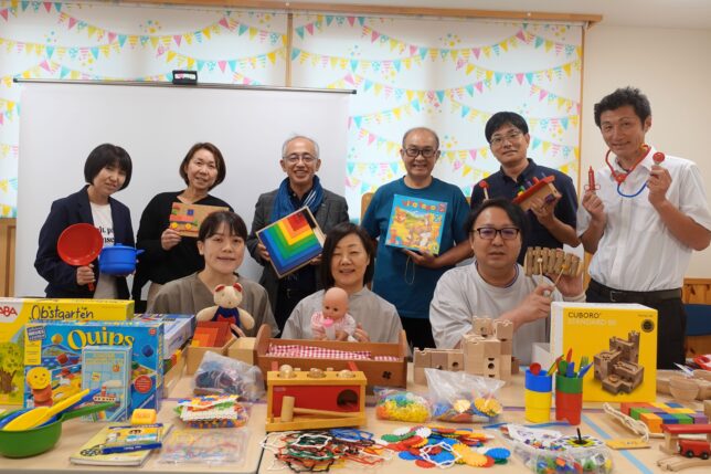 保育園の園長先生方を対象とした「おもちゃと子どもの発達」研修の集合写真。木のおもちゃを手にした参加者と講師陣が笑顔で並んでいる