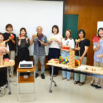 研修を終えた参加者たちの集合写真。積木、だるま落とし、木製の人形など様々な知育玩具を手に持ち、講師と一緒に記念撮影している