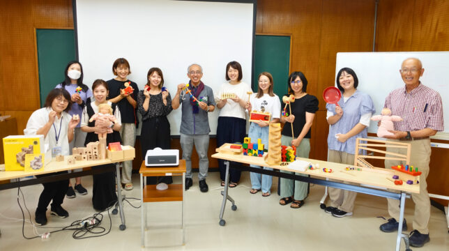 研修を終えた参加者たちの集合写真。積木、だるま落とし、木製の人形など様々な知育玩具を手に持ち、講師と一緒に記念撮影している