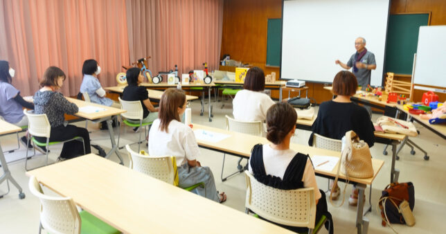 木のおもちゃを使った研修の講義風景。講師が乗り物型の玩具を手に取りながら説明し、参加者が長机に並んで熱心に耳を傾けている